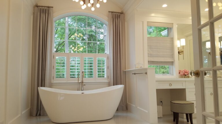 Taupe pinch pleat drapery framing an arched bathroom window with plantation shutters on the lower sash
