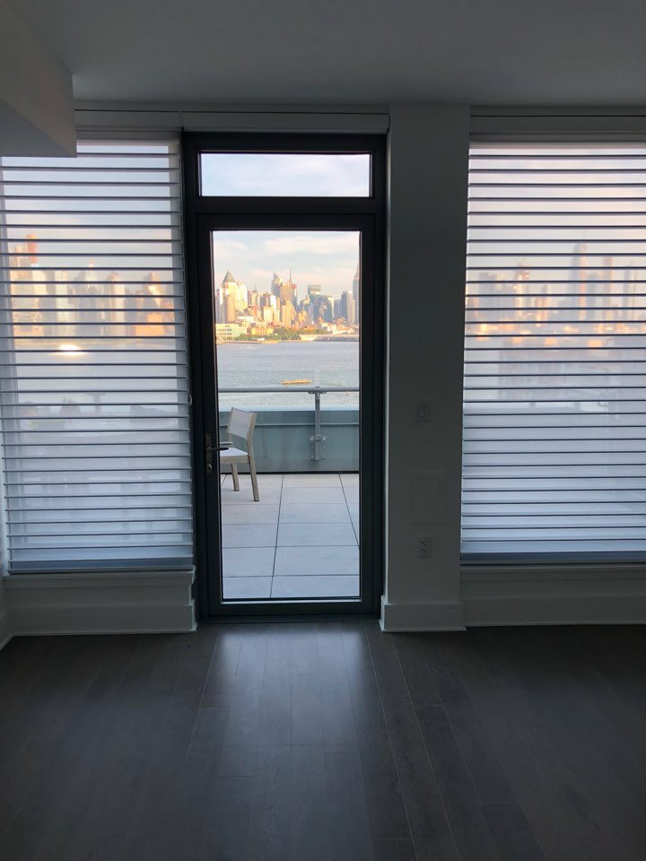 Sheer shades flanking a glass terrace door in an empty room with dark wood floors, looking over water toward a city skyline at low sun
