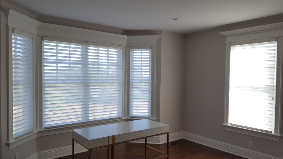 Sheer shades across a three-sided bay and a single side window in an unfurnished grey room with a small white desk on dark hardwood