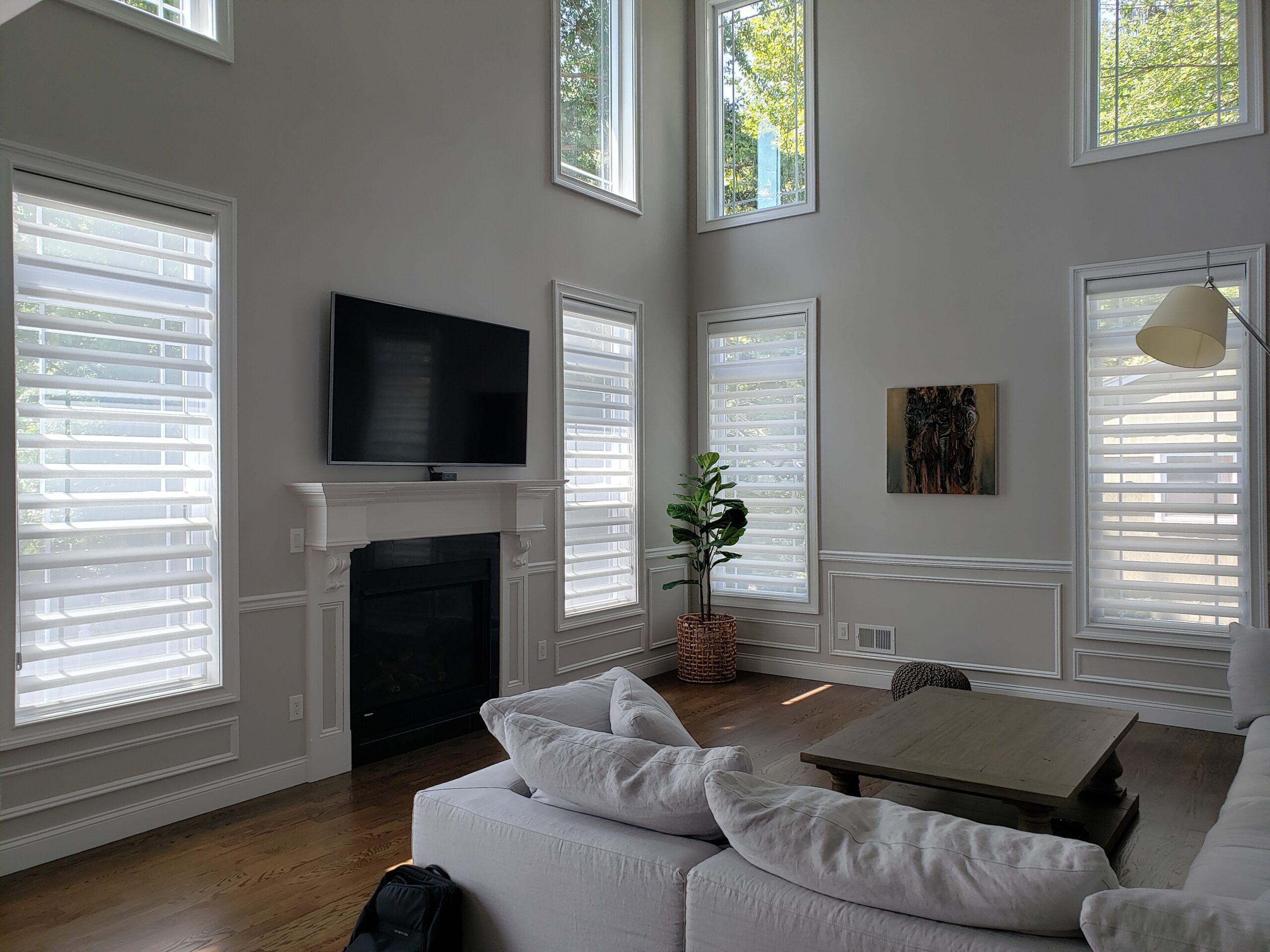 Sheer shades across tall windows and upper transoms in a two-story greige room with white wainscoting, a white fireplace mantel and a wall-mounted TV