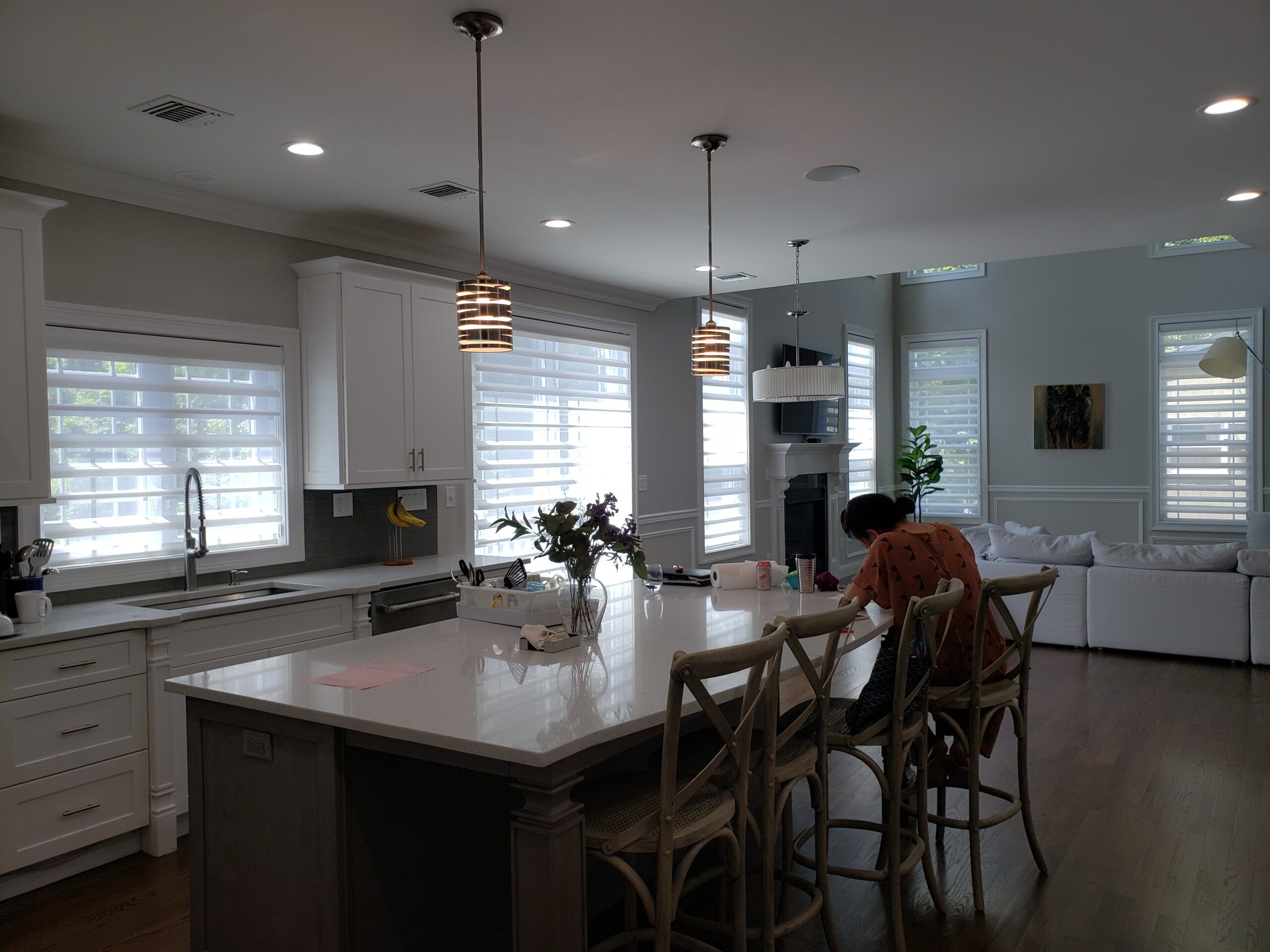 Sheer shades across a run of windows in a white open-plan kitchen with a large island and stools, opening to a gray living room with hardwood floors