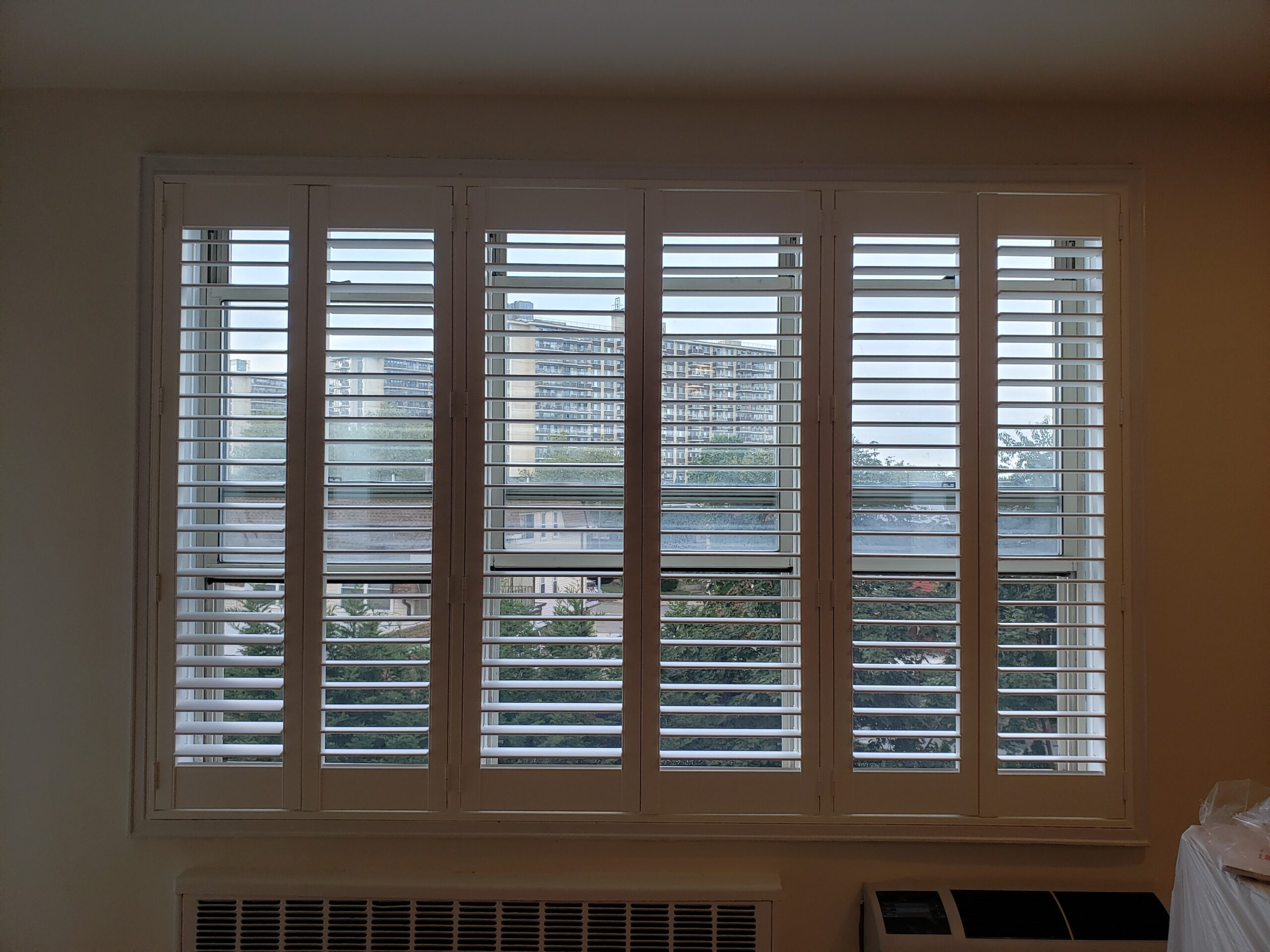 Shutters filling a wide window opening above a through-wall air conditioner in a beige-walled room, white finish, overlooking evergreens and a mid-rise apartment building
