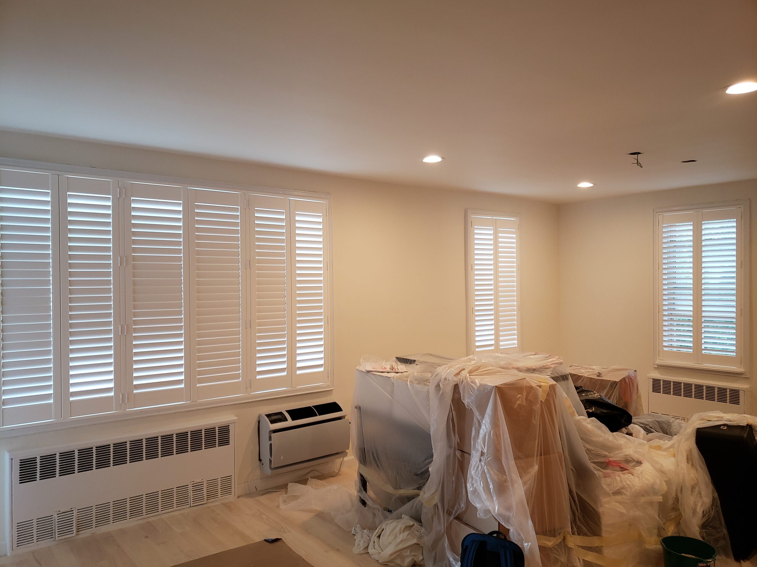 Shutters along a bank of white-framed windows in a cream-walled apartment being renovated, with white radiator covers, a through-wall air conditioner and furniture wrapped in plastic sheeting