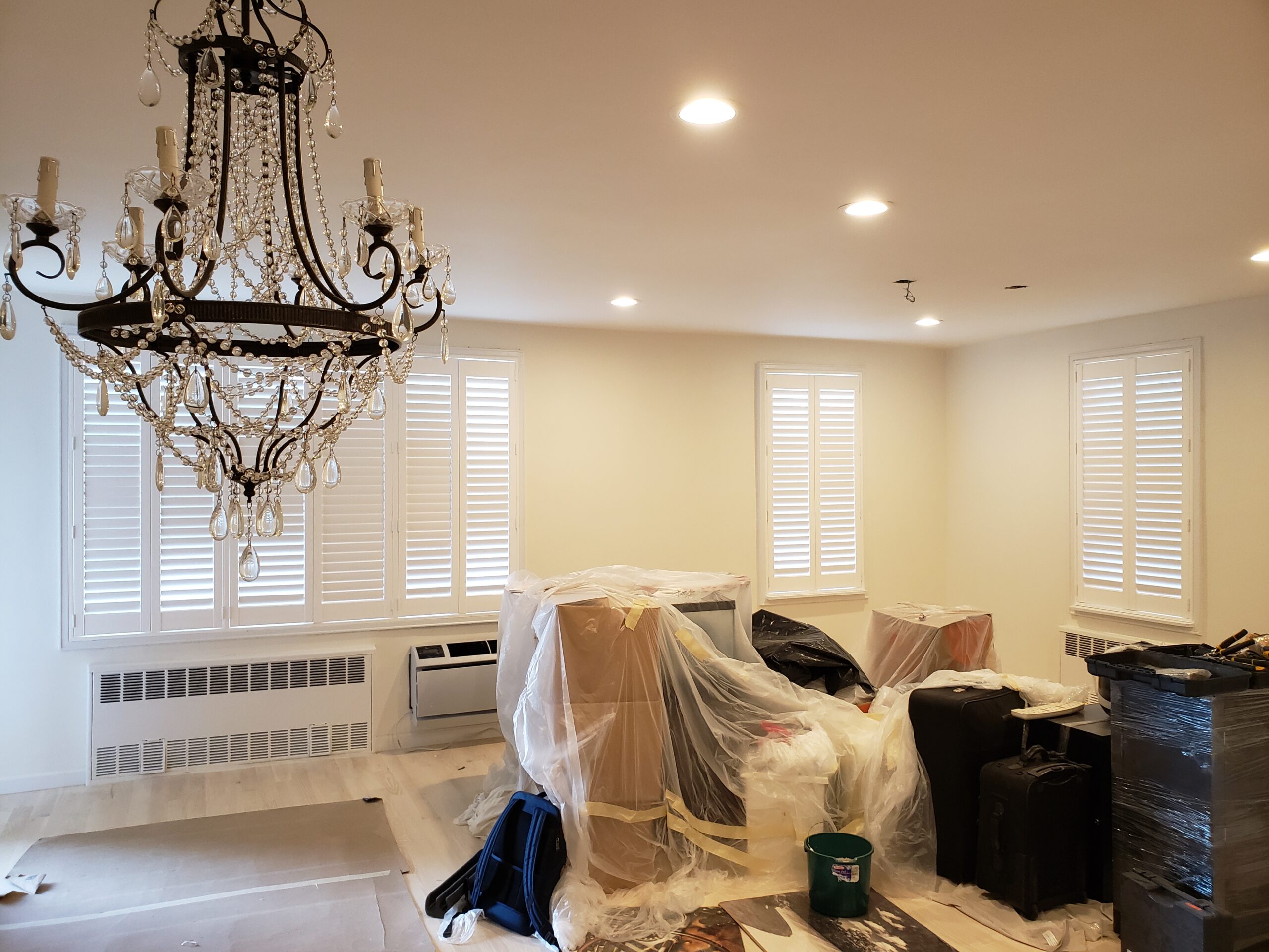 Shutters across a wall of tall windows and two narrow corner windows in a cream-walled apartment being renovated, with white radiator covers, a crystal chandelier and contents wrapped in plastic sheeting