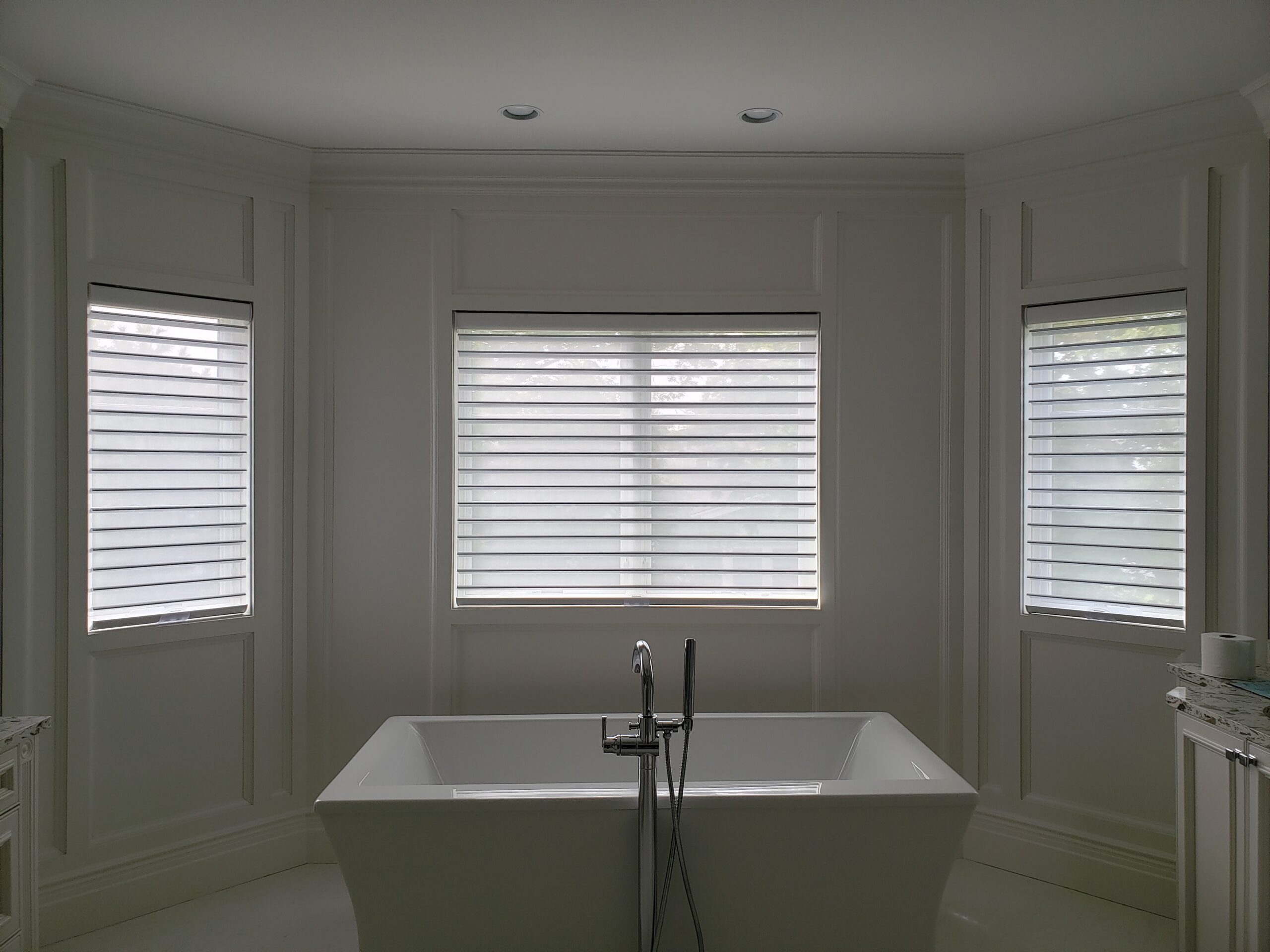 Sheer shades across three windows in a white panelled bay behind a freestanding soaking tub with a chrome floor filler