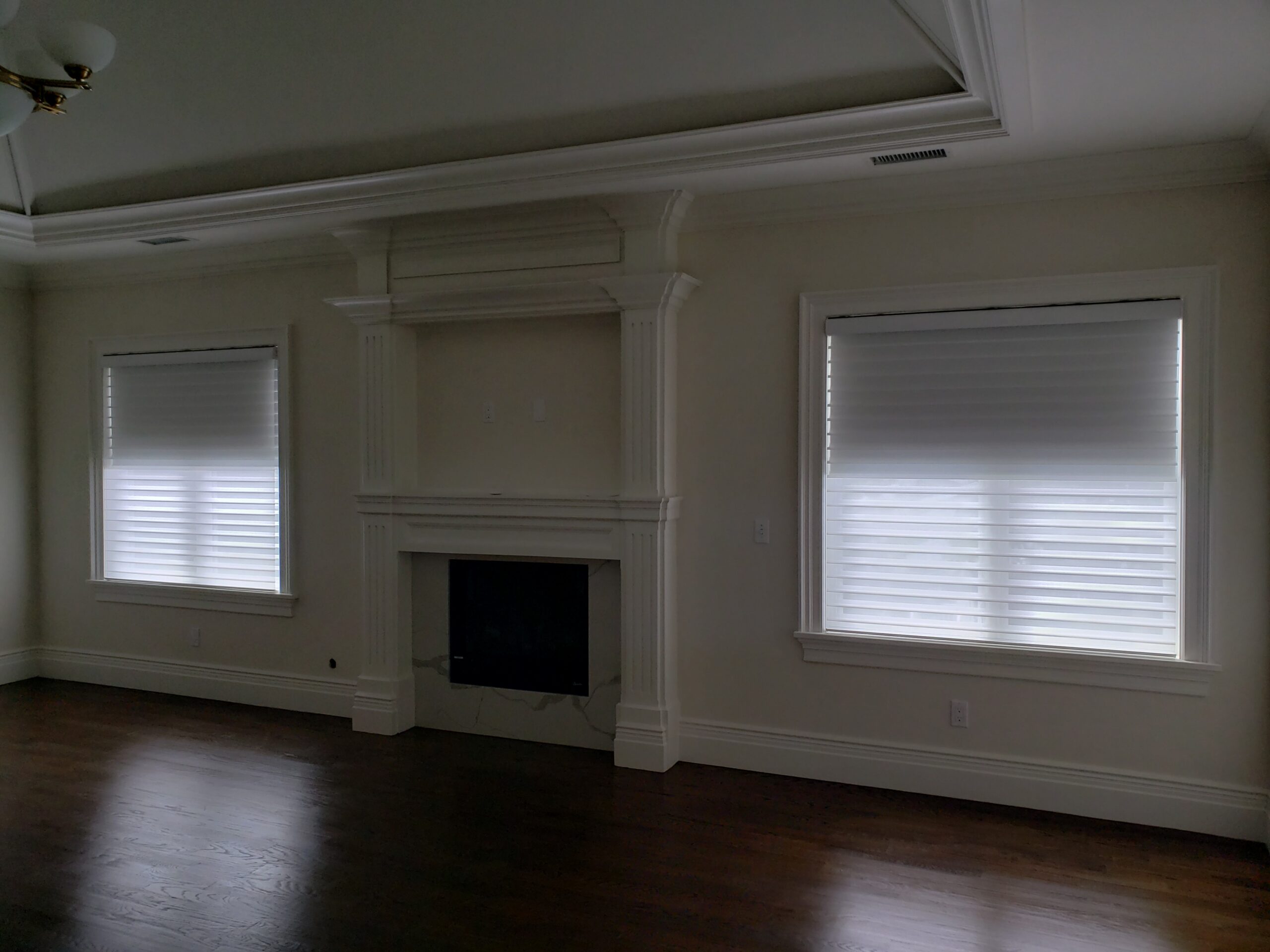 Sheer shades on two windows flanking a white painted fireplace mantel in an empty room with dark stained hardwood floors and a coffered ceiling