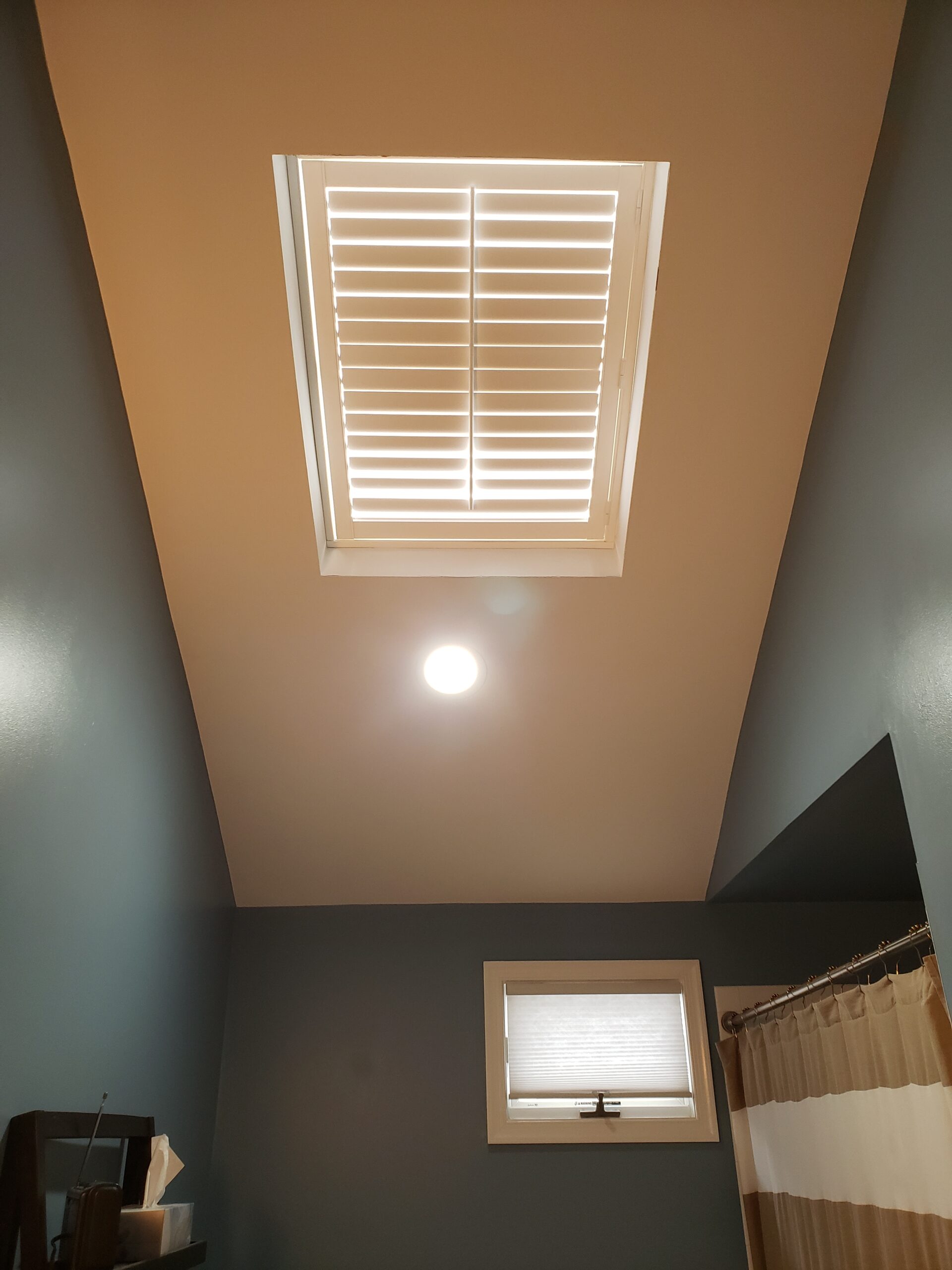 Shutters high on a tall angled wall above a striped shower curtain, with tan and slate blue paint