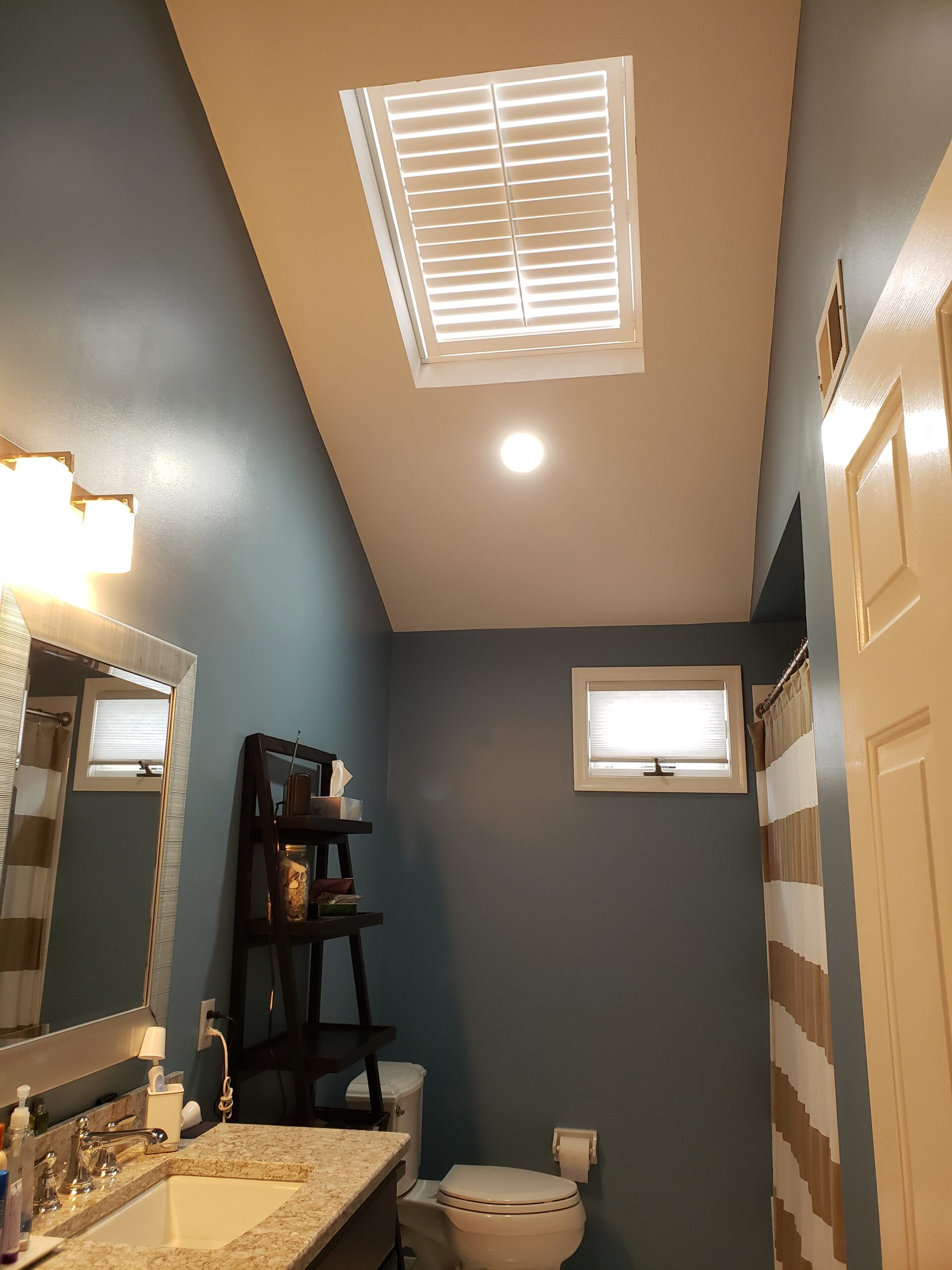 Shutters in a high angled opening on a vaulted wall of a blue-gray bathroom above the vanity and toilet