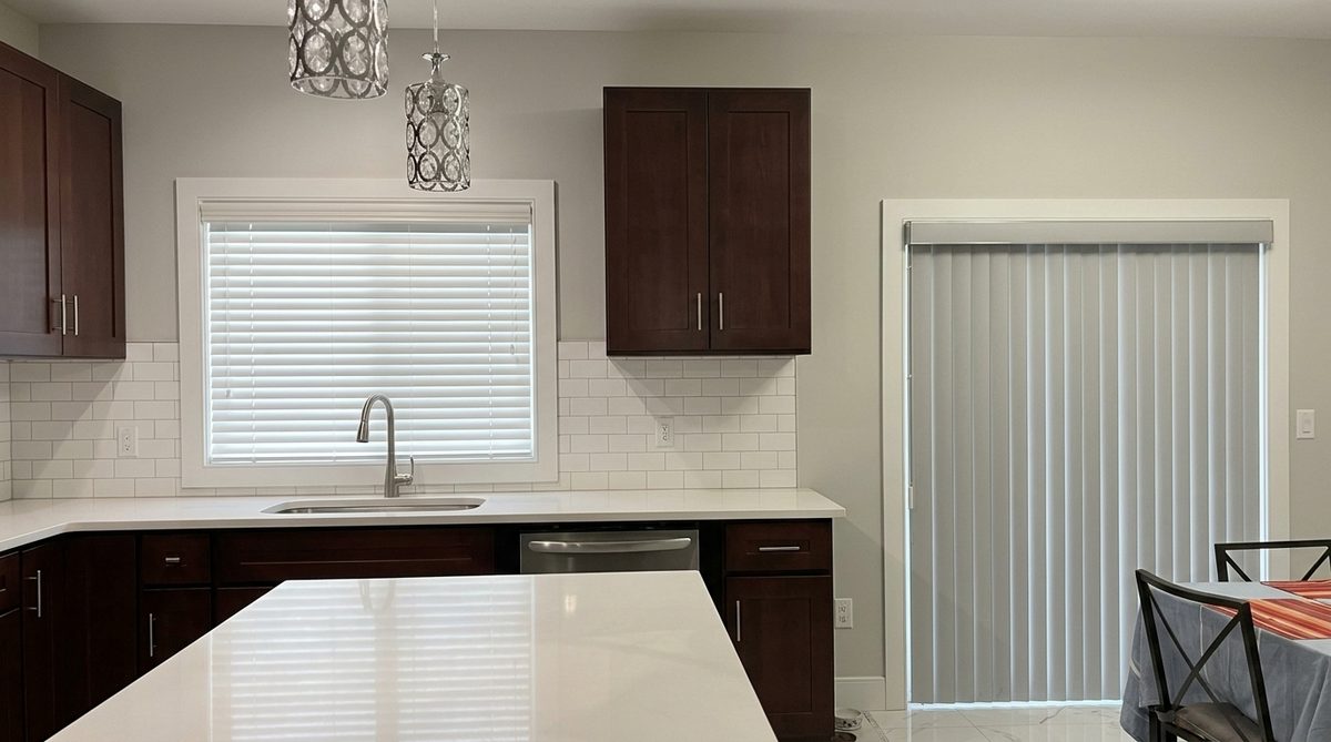 A wide white faux wood blind lowered across a large picture window in a furnished living room, slats horizontal and evenly spaced, valance across the top and a straight bottom rail